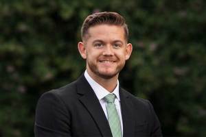 Professional headshot of Bradley Muncy wearing a dark suit, white shirt, and light green tie, standing outdoors with green foliage softly blurred in the background