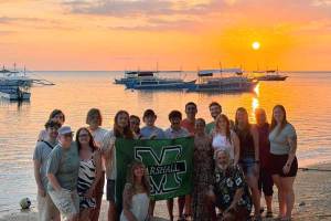 Dr. Tom Jones and Marshall University students stand on a beach holding a Marshall University flag, with small boats offshore and a colorful sunset over the water in the background