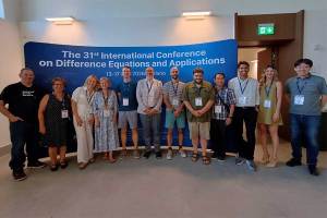 Attendees at the 31st International Conference on Difference Equations and Applications pose for a group photo in front of a conference display in Milan, Italy. Marshall University assistant professor of mathematics Dr. Tom Cuchta, wearing a green shirt and khaki shorts, stands near the center of the group.