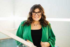 Portrait of Marshall University mathematics student Piper Cook standing beside an indoor railing, wearing a green blazer and glasses, with a bright, modern campus interior in the background