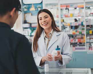 Pharmacy setting showing a pharmacist behind a counter holding a prescription bottle while assisting a patient