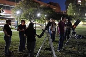 Marshall University Astronomy Club members gather on Buskirk Field at night with several telescopes set up for stargazing outside the Science Building. Trees and campus buildings are illuminated in the background as students prepare to observe the night sky.