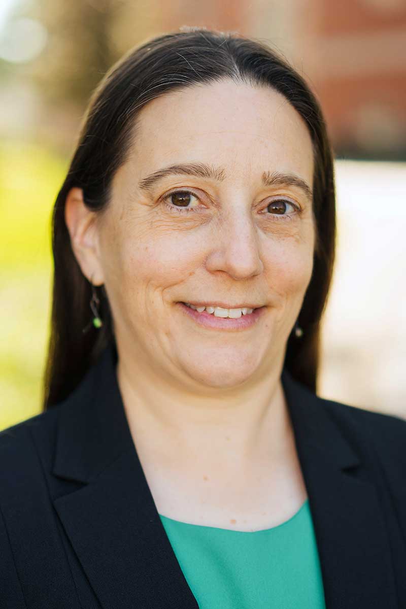 Professor of Mathematics Anna Mummert poses for a headshot outdoors, wearing a dark blazer and green top with a blurred campus background.