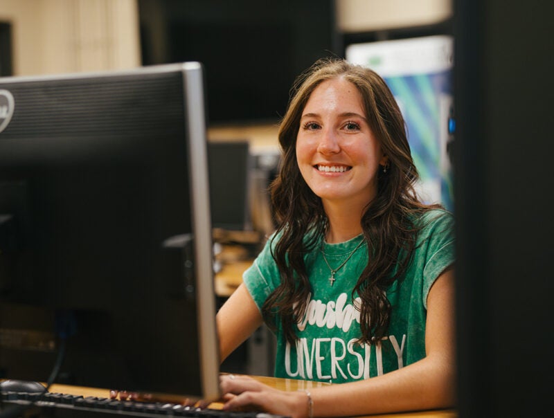 A student wearing a green, Marshall shirt smiles while working in a computer lab