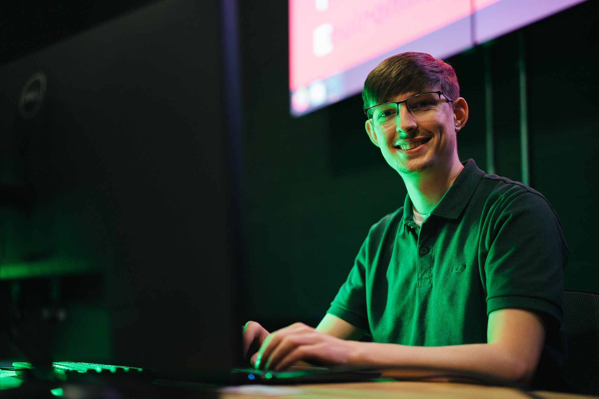 A man smiles at the camera while working on a computer in a dark lab lit by the computer screen