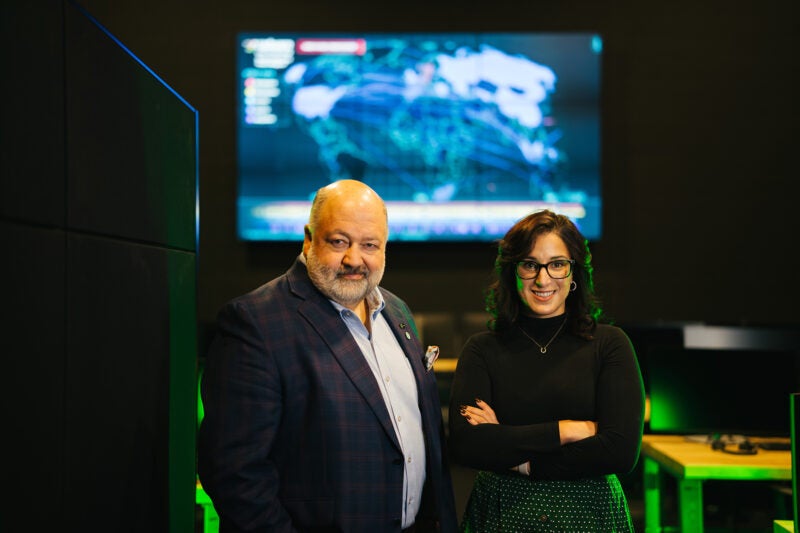 Two people stand with their arms crossed with a computer lab and large screen behind them.