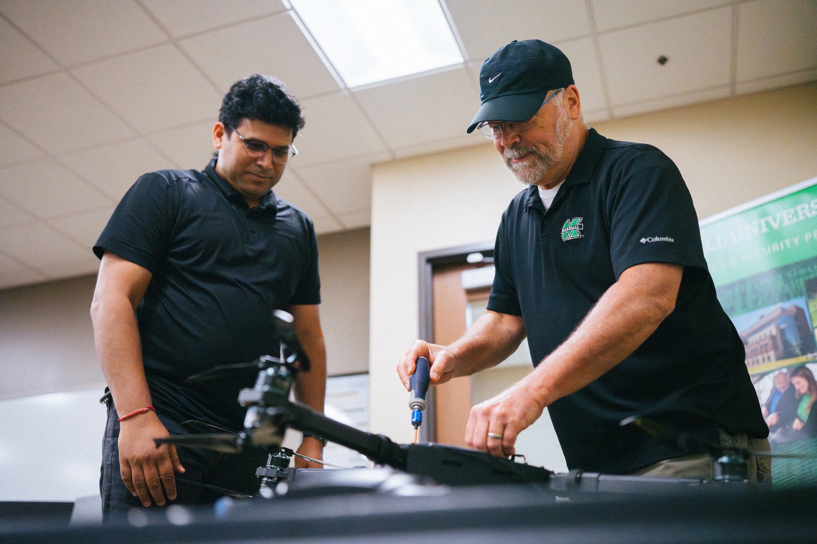 Two men work on a drone with a Marshall University Cyber security sign in the background