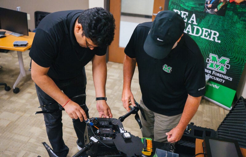 Two men work on a drone with a Marshall University Cyber security sign in the background