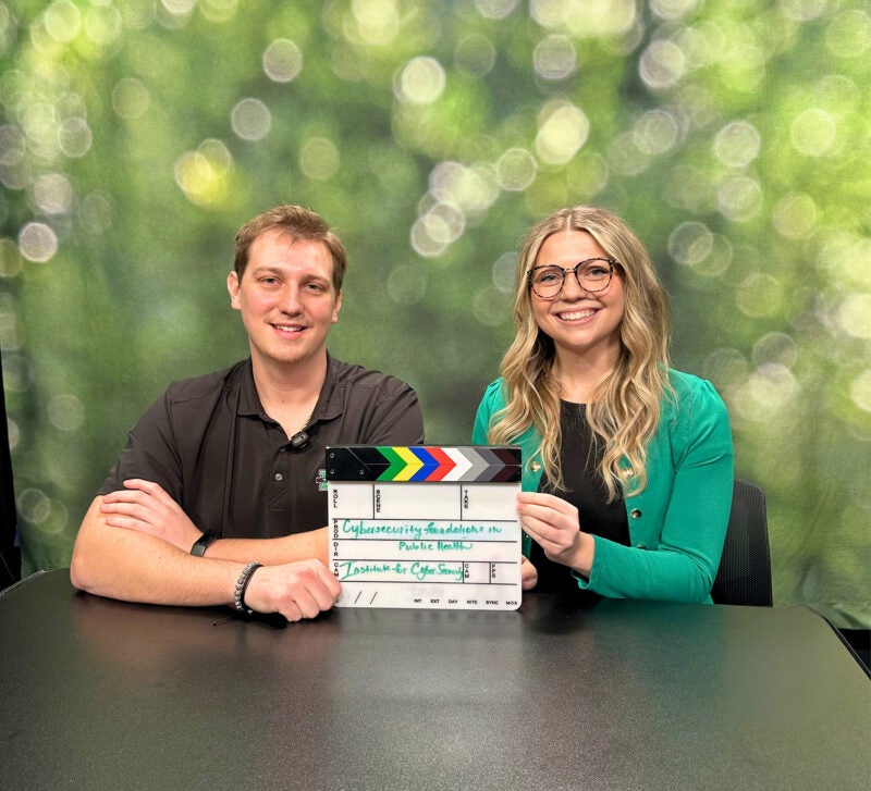 wo people sit at a table, smiling and hold a scene clapperboard in front of a sparkly, green background.