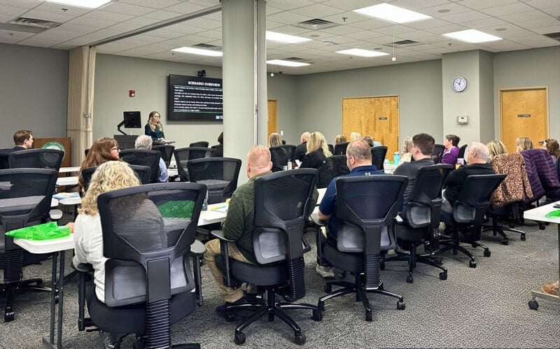 A woman conducts a training session to a room full of people sitting in back chairs at long tables.