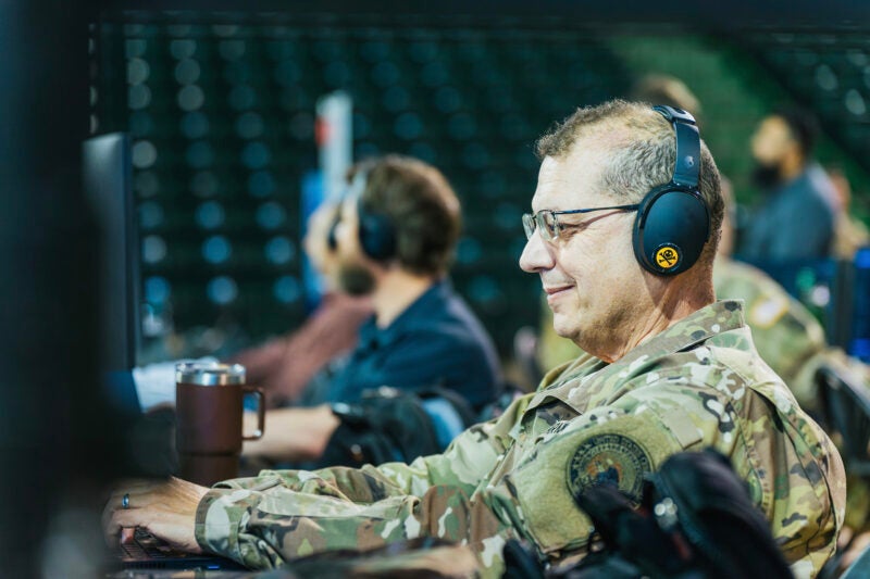 A military service member in camouflage uniform and headphones works at a computer workstation during a Lock Shield exercise, other participants are blurred in the background.