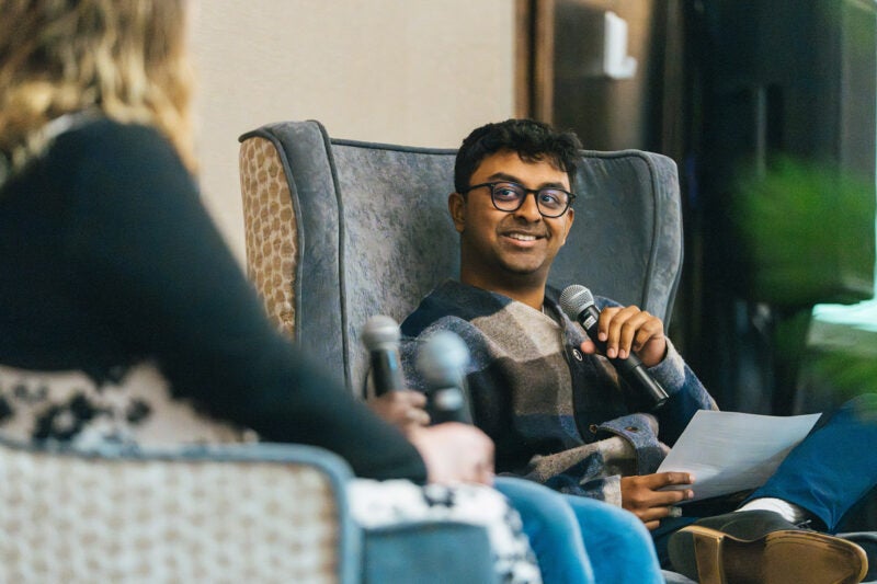 Man wearing glasses smiles while speaking into a microphone during a panel discussion, holding notes as another panelist sits in the foreground.