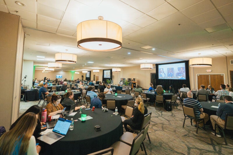 Attendees seated at round tables watch a panel discussion onstage during CyberCon, with a large projection screen.