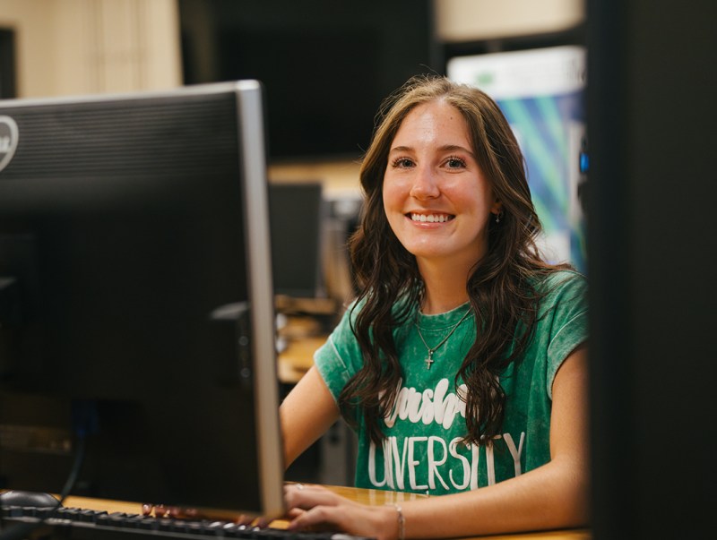 A student wearing a green, Marshall shirt smiles while working in a computer lab
