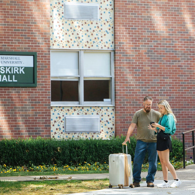 A student shows her father her phone as they stand outside of Buskirk Hall with a suitcase on Marshall University's move-in day.