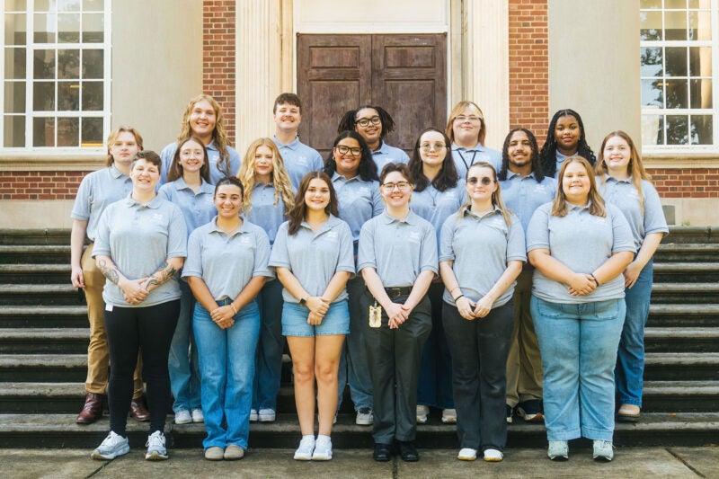 Marshall Commons staff members wear matching gray shirts and smile together on the steps of Morrow Library