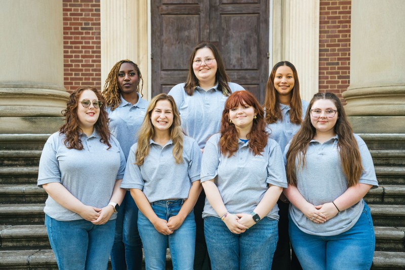 Buskirk Hall staff members wear matching gray shirts and smile together on the steps of Morrow Library