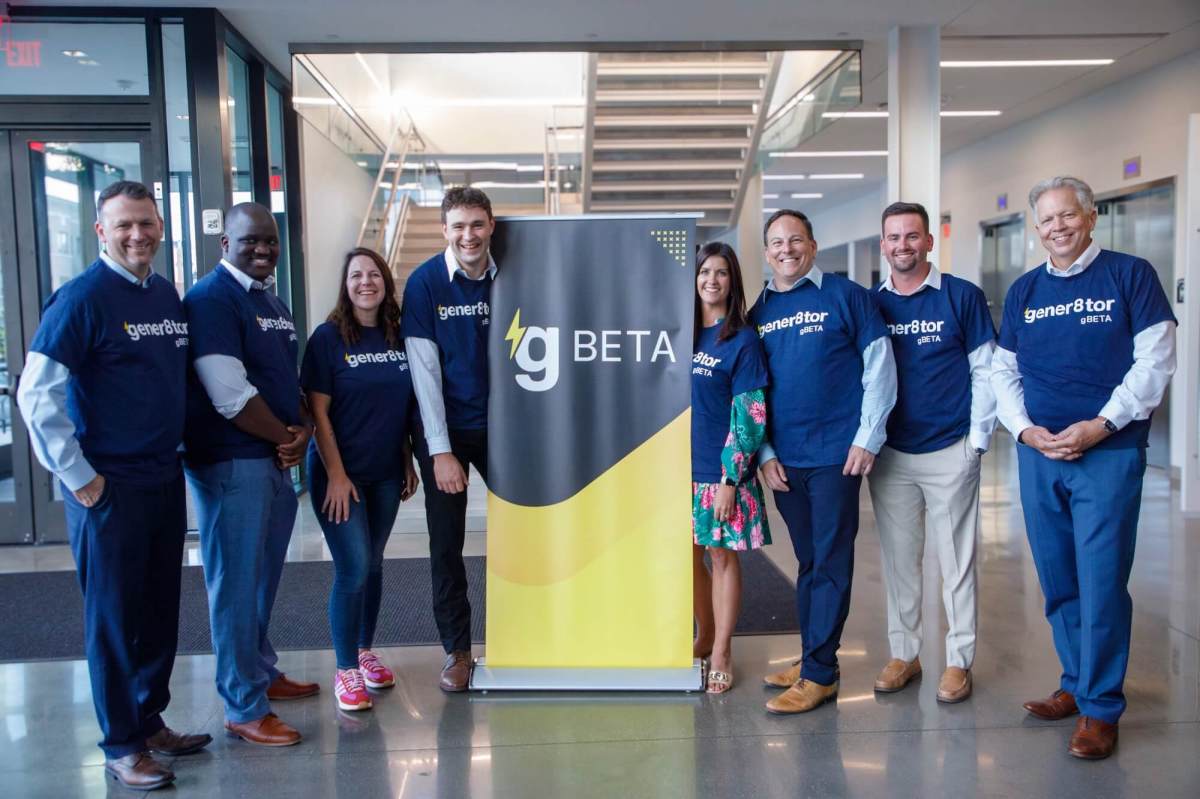 Group photo of eight people standing in a modern office building lobby around a gBETA promotional banner. The group is wearing matching navy blue gBETA t-shirts over business-casual attire and smiling at the camera. A staircase and glass-walled offices are visible in the background, with the gBETA logo prominently displayed on the banner in the center of the image.