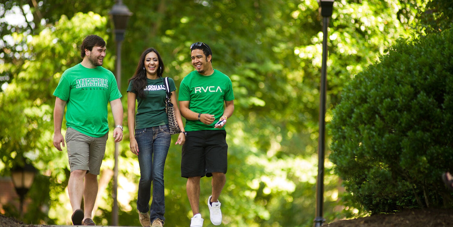 Students wearing green, Marshall University shirts walk together on a sidewalk surrounded by trees