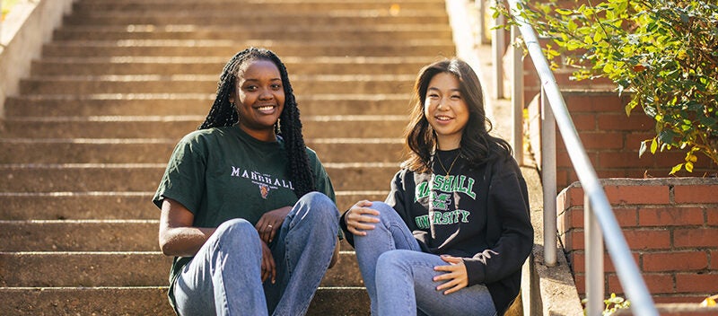 Two students smile at the camera while sitting together on concrete steps