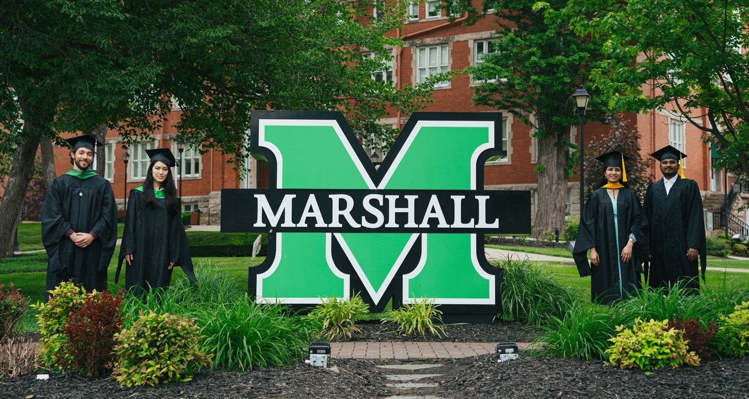 Four Marshall University students stand together wearing graduation caps and gowns with a large Marshall sign on the university's Huntington campus
