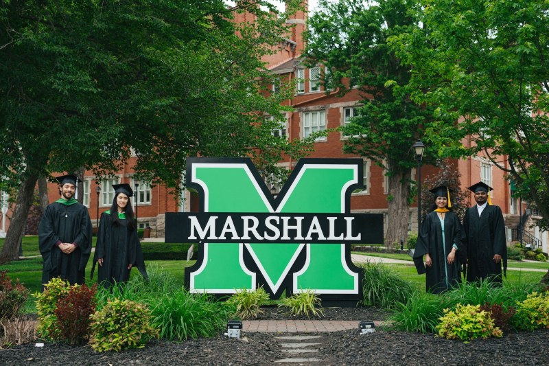 four international students standing by the block M on campus in their caps and gowns