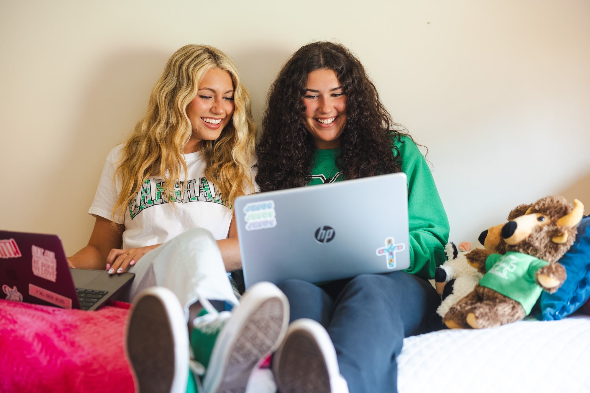 two students on a bed in a residence hall sitting up and looking at a laptop