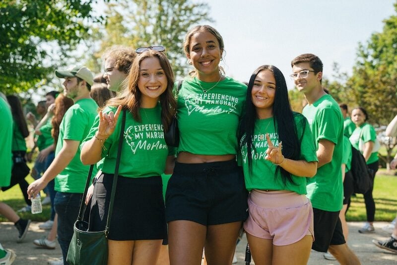 Group of students wearing green Marshall shirts smiling together outdoors on campus during an event.