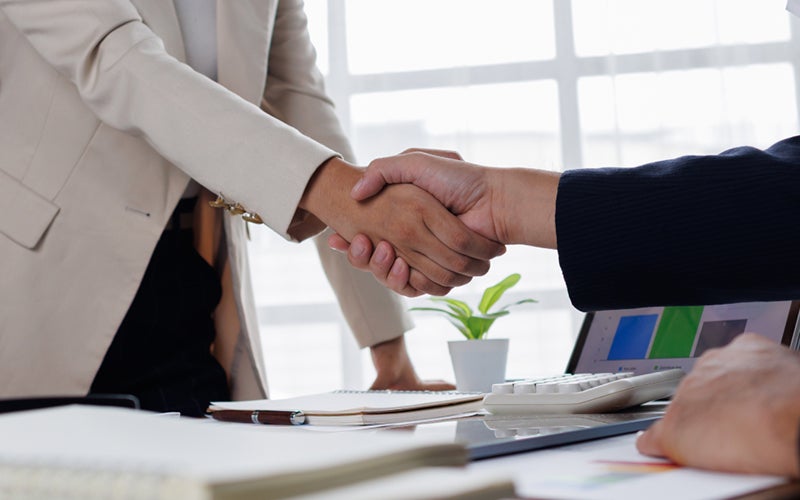 Two people shake hands over a desk covered in stacks of legal papers.