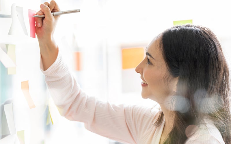 A person writes on a post-it note that is stuck to a wall.