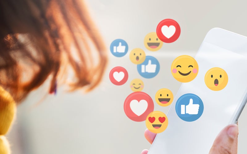 A graphic of a person holding a phone with various heart, smile and like emojis overtop