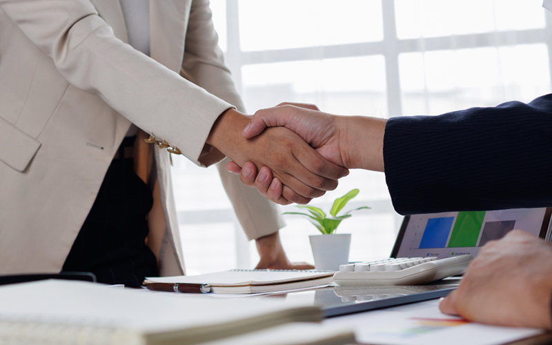 Two people shake hands over a desk covered in stacks of legal papers.