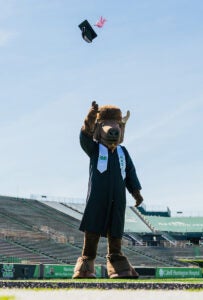 Marco, wearing a graduation gown and stole, tosses a graduation cap in Joan C Edwards Stadium