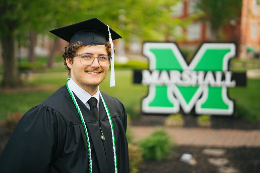 Luke Jeffery smiles while wearing his cap and gown in from of a Marshall sign at Marshall University