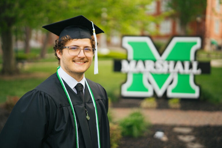 Luke Jeffery smiles while wearing his cap and gown in from of a Marshall sign at Marshall University
