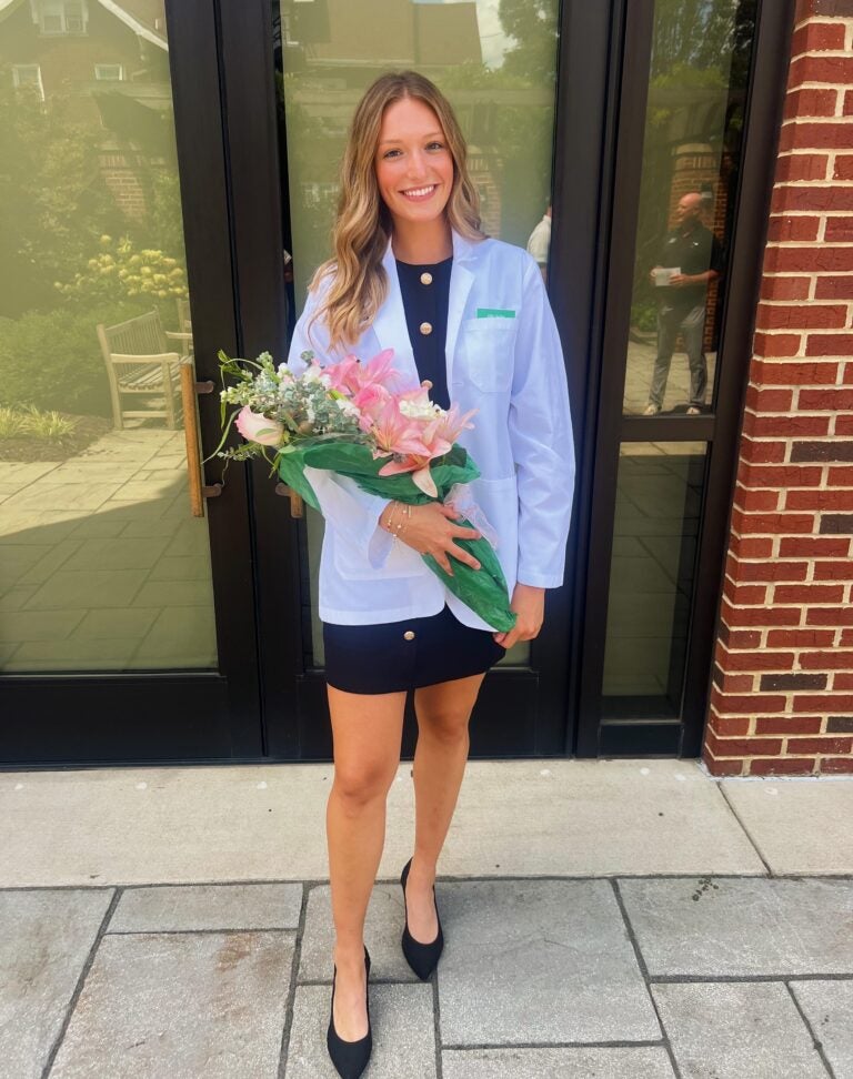 Allie Ingles stands smiling, holding a bouquet of flowers, while wearing her School of Pharmacy white coat.