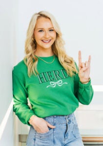 Bethany Ledford wears a green Marshall University sweatshirt while smiling and holding up bison horns with her hand 