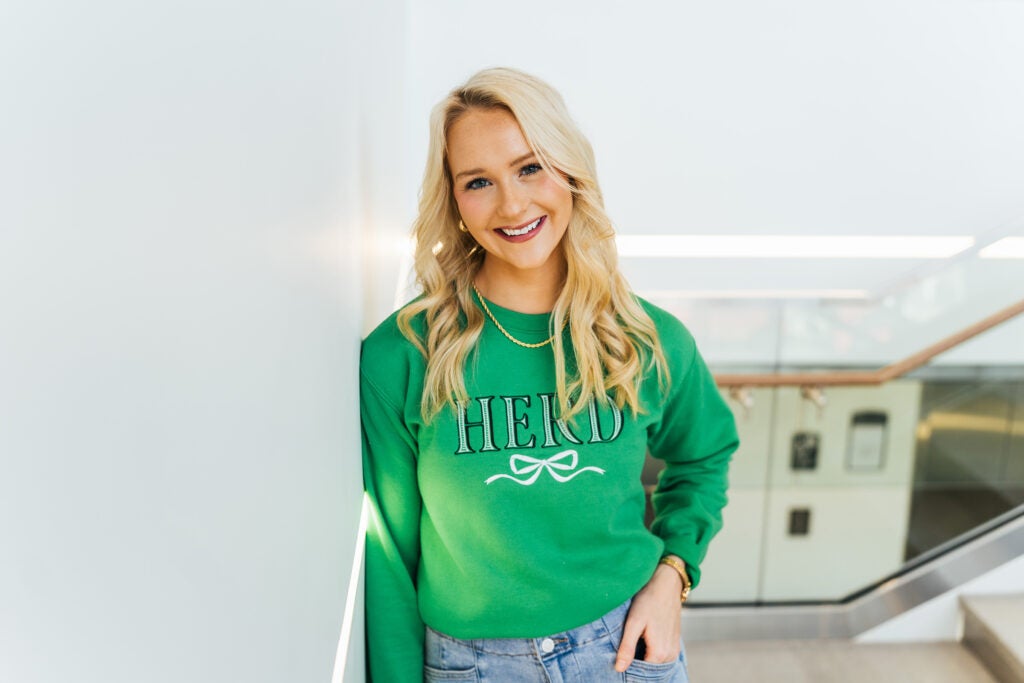 Bethany Ledford wears a green Marshall University sweatshirt while leaning against a wall inside Marshall's Brad D. Smith Center for Business and Innovation.