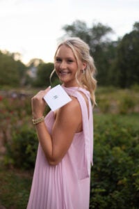 Bethany Ledford wears a pink dress and holds and invitation over her shoulder 