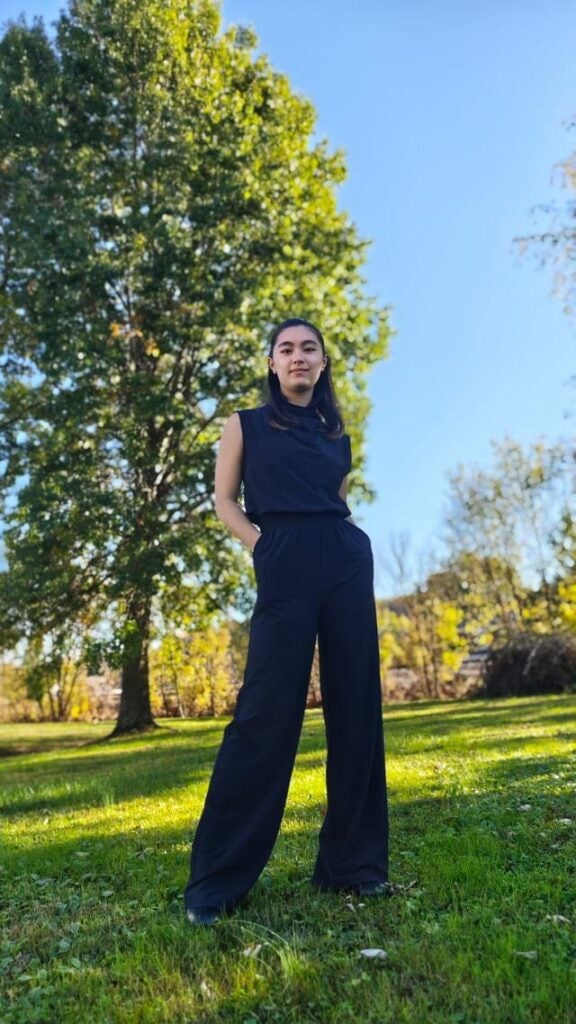 Sarah Talkington wears a black jumpsuit and smiles at the camera with a large tree behind her