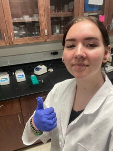 Kayleigh Garcia wears a white coat and rubber gloves giving a thumbs up while standing in a laboratory
