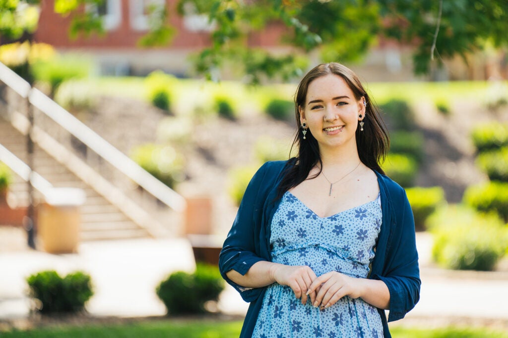 Kayleigh Garcia wears a blue floral dress and sweater and stands smiling on Marshall University's Huntington campus