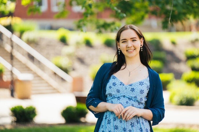 Kayleigh Garcia wears a blue floral dress and sweater and stands smiling on Marshall University's Huntington campus