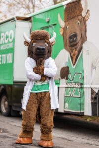 Marshall University's Marco wears a white coat and stands in front of his photo on the new medical mission truck. 