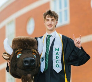 Jack Dowling wears his graduation regalia and hold's Marco's mascot head while smiling for a photo