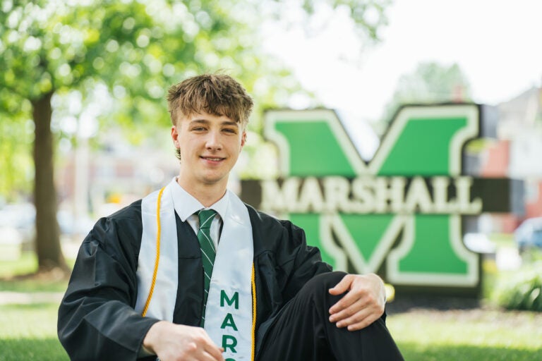 Jack Dowling wears his graduation regalia and Marco stole while sitting in front of a large Marshall sign.