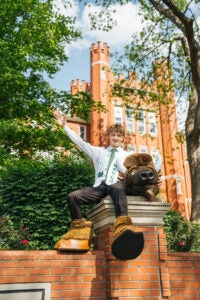 Jack Dowling wears his graduation regalia and Marco's mascot hooves while sitting next to the costume head sitting on the sign in front of Marshall's Old Main