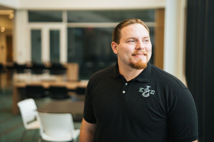 Dylan Glasscock wears a black polo shirt with Greek letters while looking out a window in a bright, modern academic building.
