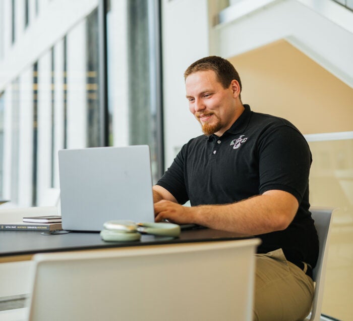 Dylan Glasscock wears a black polo shirt with Greek letters while working on a laptop computer in a bright, modern building.