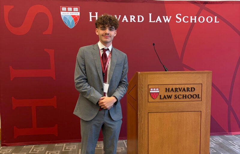 Mark Hysell wears a suit and smiles next to a Harvard Law School podium in front of a Harvard backdrop